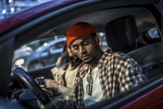 Young man wearing a beanie and plaid shirt sitting in a parked car with a woman, looking down with