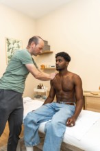 Physical therapist massaging a young black man's shoulder while he sits on an examination table,