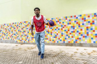 Young black man in casual sportswear playing and dribbling a basketball on a paved court against a