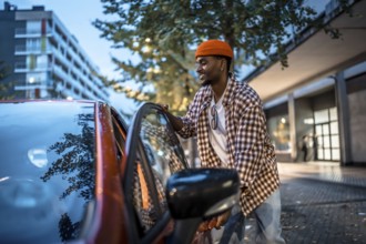 Young black man with a cheerful expression standing by an orange car, opening the door while