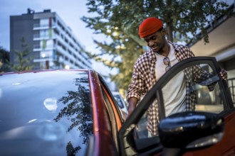 Young black man in a red beanie smiling as he opens a car door on a city street, ready to drive