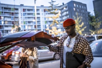Young man wearing a red beanie smiling while opening a car trunk in an urban parking lot, preparing