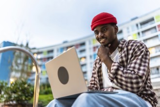 Young black man smiling while working on a laptop computer with casual clothes and a red beanie,
