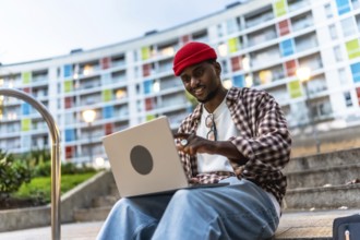 Young black man in casual clothing using a laptop while sitting on steps in an urban setting,