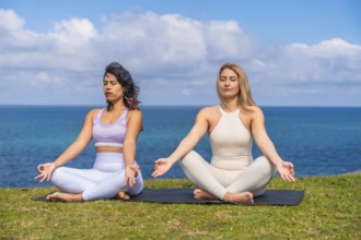Two women sit cross legged on a mat by the ocean, practicing yoga and meditation together at