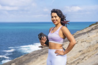 Woman smiling and holding a rolled yoga mat, standing confidently on a coastal cliff overlooking