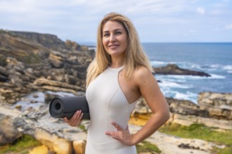 Woman stands on rocky coast by the ocean holding a yoga mat, smiling at camera before a yoga and
