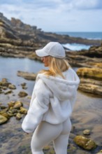 Woman in comfortable hoodie and leggings meditating on rocky coast, back to camera, finding calm