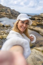 Blonde woman wearing a white cap and hoodie reaching out to take a selfie, smiling to the camera