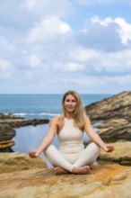 Woman in a white outfit sitting in a lotus position on a rock by the ocean, meditating and finding