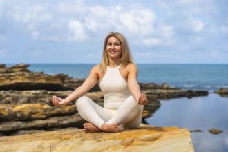 Woman practicing yoga in lotus on a rocky ocean cliff, meditating with eyes closed, embracing