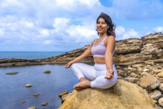 Woman practicing lotus yoga on a coastal rock, meditating peacefully at the sea's edge, embracing