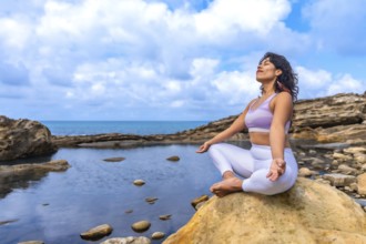 Woman in sportswear practicing yoga and meditation in lotus position on a large rock by the sea,