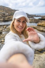 Blonde woman in white cap and jacket smiling at camera on a rocky beach, forming a heart with her