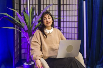 Woman sitting in a professional content creation studio, holding a laptop in her lap while looking