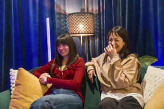 Two young women sitting on a sofa, one smiling and the other laughing, collaborating creatively in