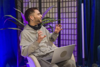 Young man wearing headphones and using a laptop, actively gesturing while recording a podcast or