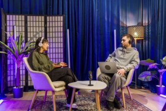 Two young adult content creators, a man and a woman, wearing headphones and sitting in chairs, are