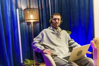 Young man wearing headphones and hoodie, sitting comfortably with a laptop in a home studio,
