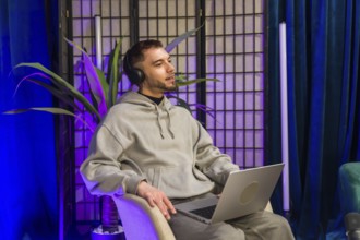 Young man wearing headphones and holding a laptop, talking and recording content for a podcast or