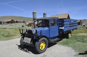 A blue historic truck with yellow wheels, Dodge Graham 1927, stands in front of a wooden house