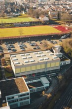 Overview of a sports field with parking lot and school complex in urban and natural surroundings in