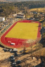 View of a large sports field with green field and red track, surrounded by cars and nature in