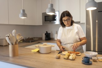 Mature woman wearing apron cutting carrots and potatoes while cooking at home in a modern kitchen.