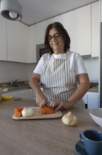 Mature woman wearing apron cutting carrots on wooden cutting board in modern kitchen. Healthy