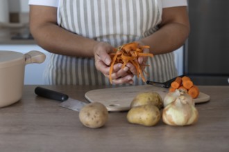 Close-up view of woman's hands holding carrot peels while peeling fresh carrots on a wooden board.