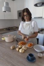 Woman wearing apron preparing vegetables for homemade soup while peeling carrots in modern kitchen.