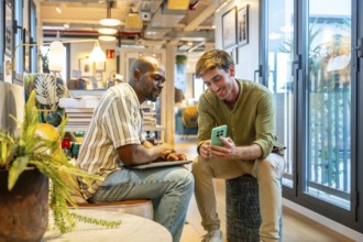 Two diverse male colleagues are sharing information on a mobile phone and laptop while working