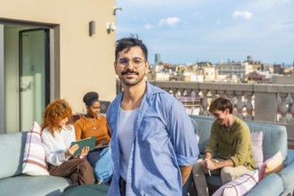 Smiling man standing on a rooftop terrace as diverse colleagues relax and work in the background,