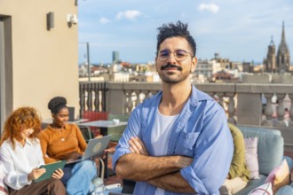 Young man with crossed arms and confident smile while diverse colleagues collaborate and chat