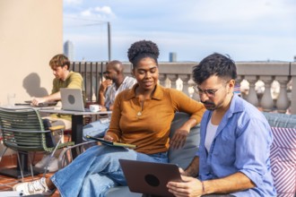 Diverse colleagues working and interacting on a rooftop terrace, using laptops and tablets while