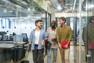 Three diverse male colleagues walking through a modern office corridor, smiling and engaging in