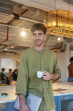 Young man standing in a modern coworking office, enjoying a coffee break while holding a laptop,