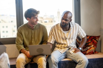 Two diverse male colleagues are sitting together, looking at a laptop screen, discussing ideas, and