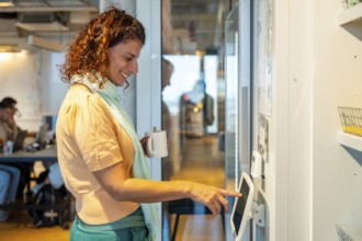 Woman standing in a modern office or coworking space, smiling and holding a coffee cup while using