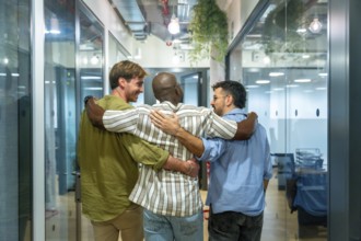 Three diverse male colleagues walk into a modern office corridor embracing and smiling, conveying