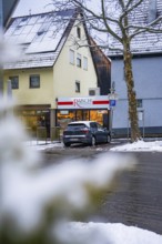 Snowy city street with parked car in front of a building with a shop, Gechingen, Calw district,