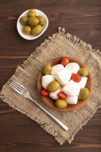 White Mozzarella cheese, with tomatoes and olives in wooden bowl on brown wooden background and