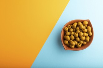 Fresh green olives in ceramic clay bowl on blue and orange pastel paper background. Top view, flat