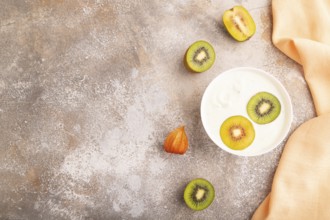 Yogurt with kiwi in white bowl on brown concrete background and orange linen textile, top view,