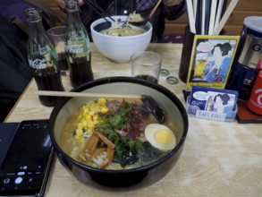 A bowl of ramen with egg, corn and vegetables on a table in a restaurant, accompanied by cola