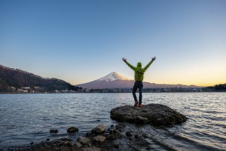 Young man stretching his arms in the air, standing on rocks in Lake Kawaguchi, view of Mount Fuji