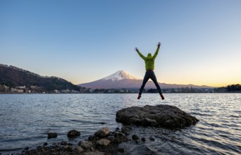 Young man jumping and stretching his arms in the air, standing on rocks in Lake Kawaguchi, view of