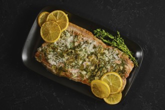 Roasted pink salmon is served on a black plate with slices of lemon and herbs. This dish is ready