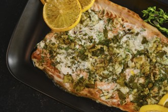 A pink salmon fillet is ready for serving on a black plate. It is garnished with herbs and slices