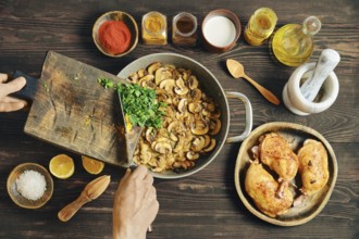 Unrecognizable person adds chopped herbs to a frying pan with fried sliced mushrooms. Nearby, bowls
